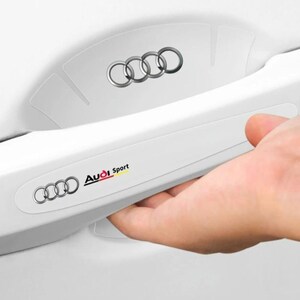 May include: White car door handle with the Audi logo and the words "Audi Sport". The handle has a silver logo on the door. A hand is reaching for the handle.