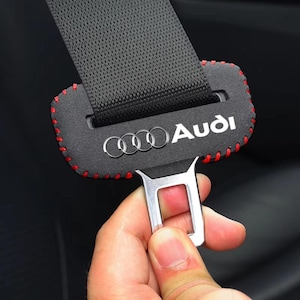 May include: Black seat belt cover with red stitching and the Audi logo in white. The seat belt is black and the buckle is silver. The cover is rectangular and wraps around the seat belt.