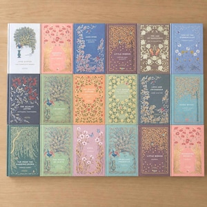 Puede incluir: Una colección de libros de tapa dura con cubiertas decorativas. Los libros presentan diseños florales y de naturaleza en varios colores, incluyendo azul, rosa y verde. Los títulos y nombres de los autores son visibles en los lomos. Los libros están ordenados sobre una superficie de madera.