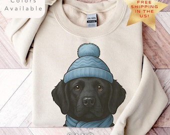 Può includere: Felpa color sabbia con un Labrador Retriever nero che indossa un cappello e una sciarpa di maglia blu chiaro. La grafica è un'illustrazione dettagliata del volto del cane. Il testo sull'immagine dice "Multiple Colors Available".