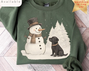 Può includere: Felpa verde militare con un pupazzo di neve con cappello a cilindro e sciarpa, e un cane nero. Il design include una scena innevata con pini. Il testo "Multiple Colors Available" è in un riquadro bianco.
