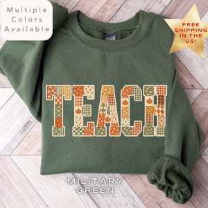 May include: A military green crewneck sweatshirt featuring the word TEACH in large block letters. Each letter is filled with a patchwork pattern of autumn-themed motifs including pumpkins, flowers, and plaid designs in orange, brown, and green.