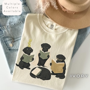 May include: An ivory t-shirt featuring a graphic of four black Labrador dogs reading books, with one dog balancing tennis balls on its head. The shirt is layered over light blue denim jeans and a tan wide-brimmed hat. Text reads Multiple Colors Available.