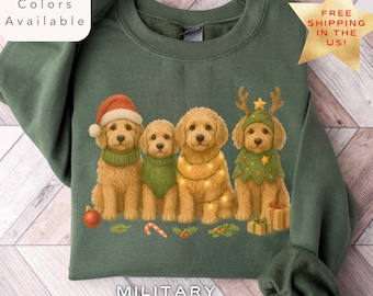 May include: Military green sweatshirt featuring a festive illustration of four golden retriever puppies dressed in holiday attire. One puppy wears a Santa hat, another a green scarf, and two are adorned with lights and a Christmas tree hat. The text "Multiple Colors Available" is visible.
