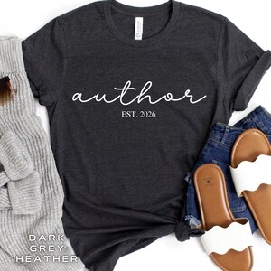 Può includere: T-shirt grigio scuro mélange con la parola "author" in corsivo bianco e il testo "EST. 2026" sotto. La maglietta è abbinata a un maglione grigio, occhiali da sole, jeans blu e sandali bianchi.
