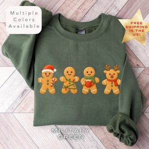Może przedstawiać: Zielona wojskowa bluza z czterema piernikowymi ludzikami w świątecznych strojach. Jeden ma czapkę Świętego Mikołaja i laskę cukrową, inny ma światełka, trzeci trzyma kubek, a ostatni szalik renifera. Tekst "Multiple Colors Available" znajduje się po lewej stronie u góry.