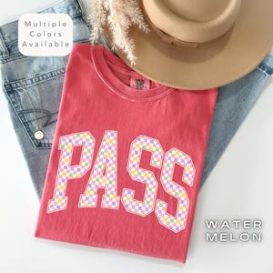 May include: A red t-shirt with the word "PASS" in a checkered pattern, paired with blue jeans and a tan hat. The shirt is labeled "WATER MELON" and "Multiple Colors Available". The shirt is a Comfort Colors brand.