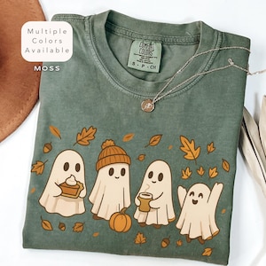以下が含まれることがあります： モスグリーンのTシャツ。漫画のゴーストのデザイン、秋の葉、カボチャが描かれています。シャツのタグには「Comfort Colors」の文字。金のネックレスがシャツにかけられています。「Multiple Colors Available」の文字が左上にあります。