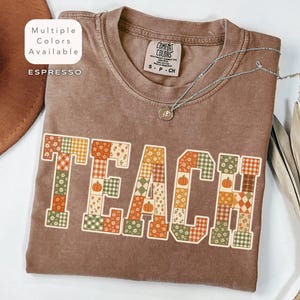 May include: An espresso-colored t-shirt with the word TEACH printed in large block letters. Each letter features a patchwork design of orange, green, and white patterns including pumpkins, flowers, and plaid. The shirt has a crew neckline.