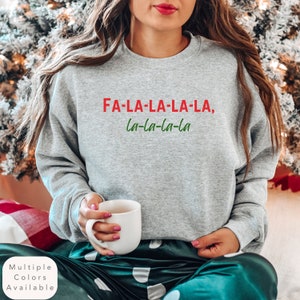 以下が含まれることがあります： 赤と緑で「FA-LA-LA-LA-LA, la-la-la-la」と書かれたグレーのスウェットシャツ。白いマグカップとグリーンの水玉模様のパンツも写っています。このスウェットシャツは、ホリデーをテーマにしたコレクションの一部です。