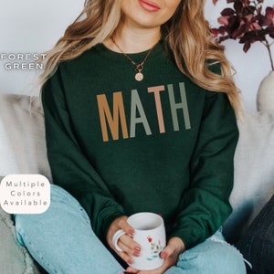May include: A forest green sweatshirt with the word "MATH" printed on the front in orange, green, and pink. The sweatshirt is being worn by a person sitting on a couch.