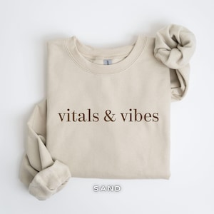 May include: A beige sweatshirt with the text "vitals & vibes" printed in brown on the front. The sweatshirt is folded and has a tag that says "SAND".