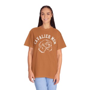 Peut inclure: Un t-shirt couleur rouille avec l'inscription "CAVALIER MOM" en blanc, en arc de cercle, au-dessus d'une illustration blanche d'un chien Cavalier King Charles Spaniel. Le t-shirt est en mati&egrave;re souple.