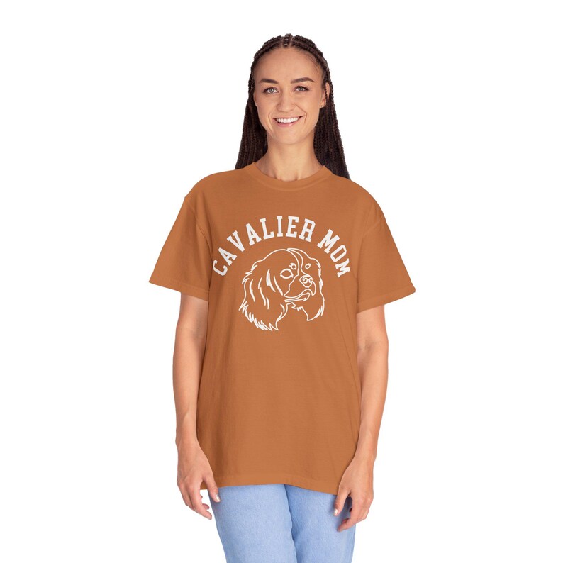 Peut inclure: Un t-shirt couleur rouille avec l'inscription "CAVALIER MOM" en blanc, en arc de cercle, au-dessus d'une illustration blanche d'un chien Cavalier King Charles Spaniel. Le t-shirt est en mati&egrave;re souple.
