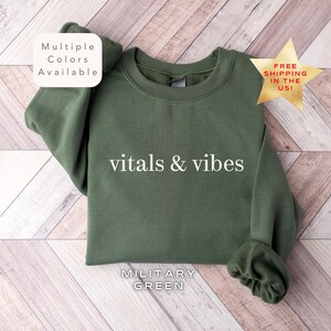 May include: A green sweatshirt with the text "vitals & vibes" printed in white. The sweatshirt is available in multiple colors and is labeled "Military Green".