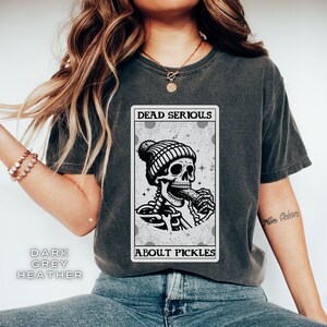 May include: A dark grey heather t-shirt with a graphic of a skeleton wearing a beanie and eating a pickle. The graphic is in a card-like frame with the text "Dead Serious About Pickles".