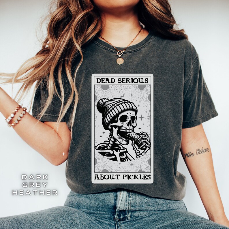 May include: A dark grey heather t-shirt with a graphic of a skeleton wearing a beanie and eating a pickle. The graphic is in a card-like frame with the text "Dead Serious About Pickles".