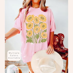 May include: A pink t-shirt with a vintage-style graphic of yellow sunflowers. The text "Multiple Colors Available" and "BLOSSOM" are printed on the bottom of the shirt.