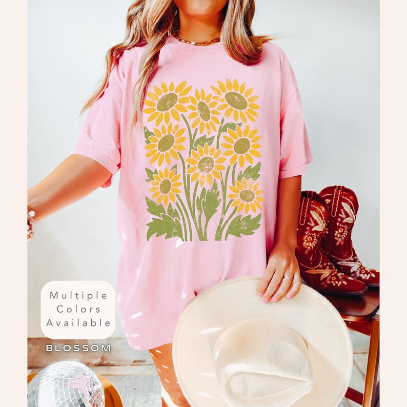 May include: A pink t-shirt with a vintage-style graphic of yellow sunflowers. The text "Multiple Colors Available" and "BLOSSOM" are printed on the bottom of the shirt.