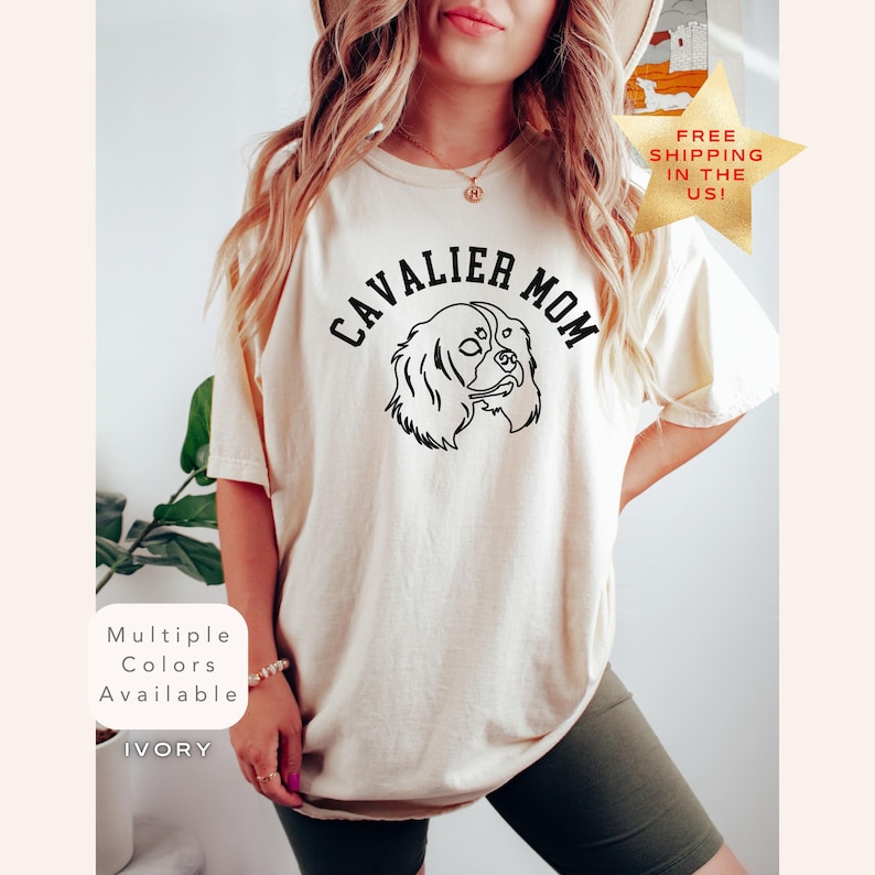 Peut inclure: T-shirt de couleur ivoire avec l'inscription "CAVALIER MOM" au-dessus d'un dessin au trait noir d'un chien Cavalier King Charles Spaniel. Le t-shirt porte &eacute;galement l'inscription "Multiple Colors Available" et "IVORY" en bas &agrave; gauche.