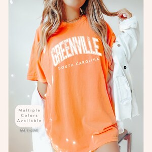 以下が含まれることがあります： 「GREENVILLE SOUTH CAROLINA」の文字が白の掠れたフォントでプリントされた、明るいオレンジ色のTシャツ。Tシャツは半袖です。「Multiple Colors Available」と色「MELON」の文字も確認できます。
