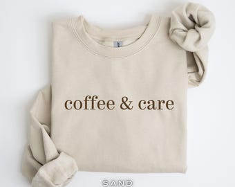 Op de afbeelding: Een beige sweatshirt met de tekst "coffee & care" in bruin gedrukt op de voorkant. Het sweatshirt is gevouwen en het label zegt "SAND".