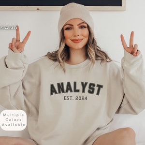 May include: A beige sweatshirt with the text "ANALYST EST. 2024" in black. The model is wearing the sweatshirt and has a peace sign with both hands.