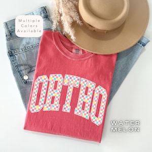 May include: A red t-shirt with the word "ORTHO" in a checkered pattern. The shirt is paired with blue jeans and a tan hat. The text "Multiple Colors Available" and "WATER MELON" are also visible.