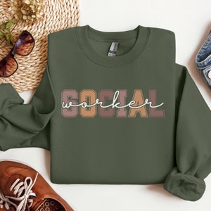 Puede incluir: Una sudadera verde con las palabras "SOCIAL WORKER" impresas en letras marrones, naranjas y rosas. Las letras tienen una fuente vintage de estilo universitario.