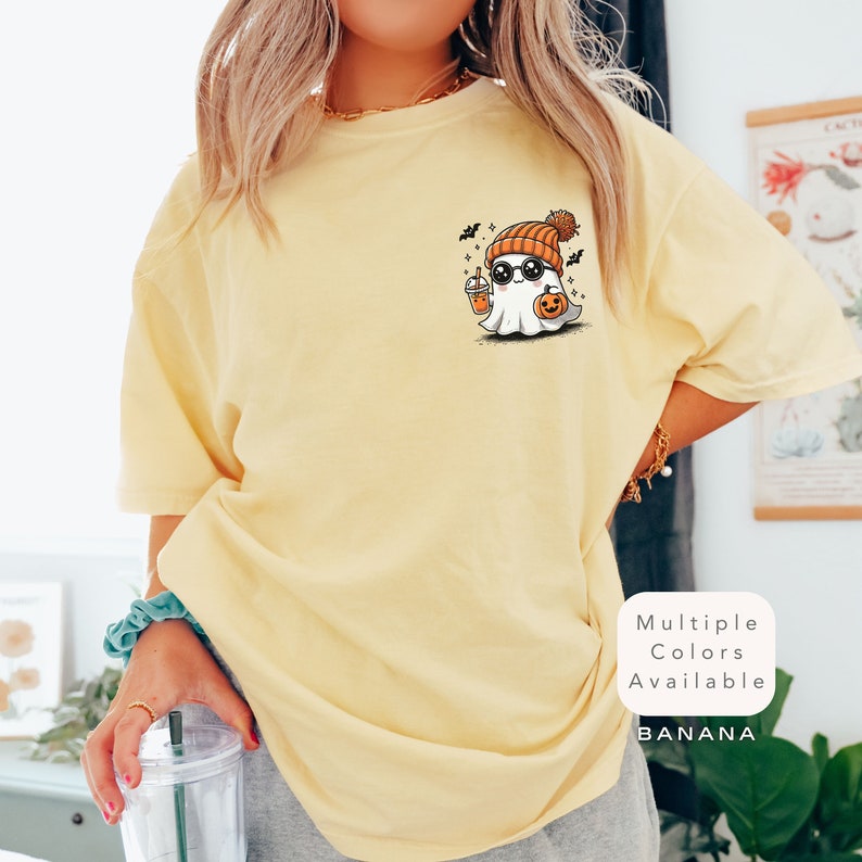 Pode incluir: Uma camiseta amarela com um fantasma de desenho animado usando um gorro e &oacute;culos de sol, segurando uma ab&oacute;bora e uma bebida. O texto "Multiple Colors Available" e "BANANA" est&atilde;o no canto inferior direito da imagem.