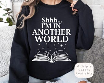May include: Black sweatshirt with white text that reads "Shhh... I'm in another world" and a white illustration of an open book with stars above it. The text "Multiple Colors Available" is in the bottom right corner.