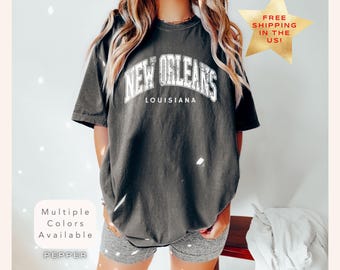 May include: A dark grey t-shirt with the text "NEW ORLEANS LOUISIANA" in white. The shirt is worn by a person. The image also includes text that says "Multiple Colours Available" and "Pepper".