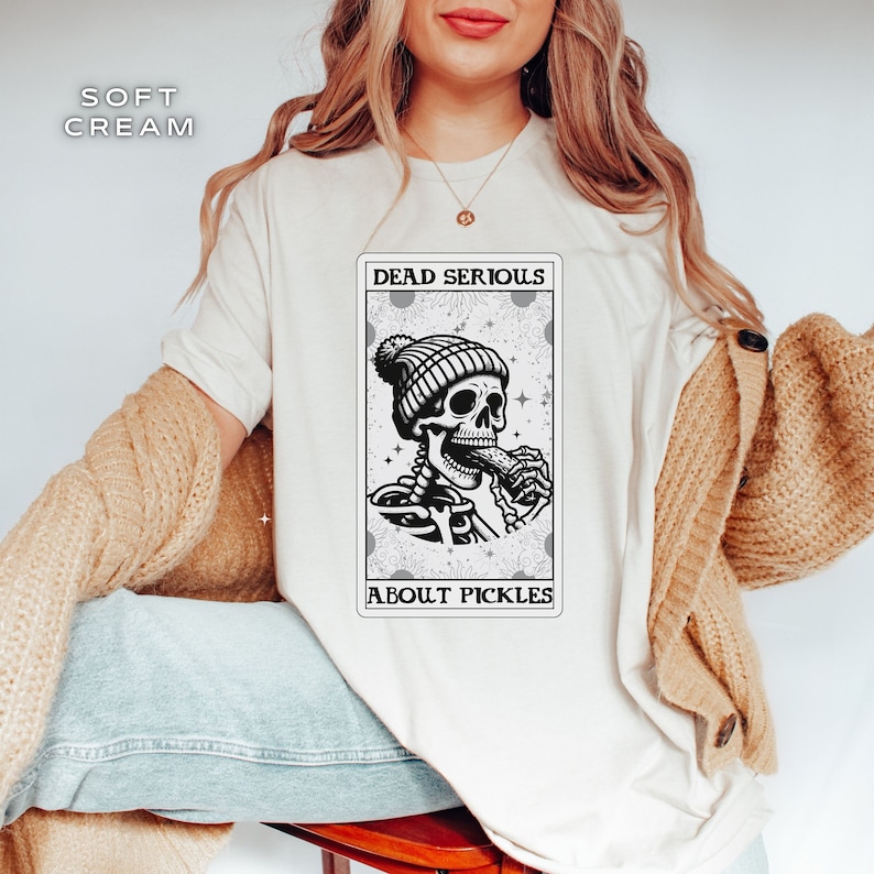 May include: A white t-shirt with a black and white illustration of a skeleton wearing a beanie and eating a pickle. The illustration is in the style of a tarot card and says "Dead Serious About Pickles".