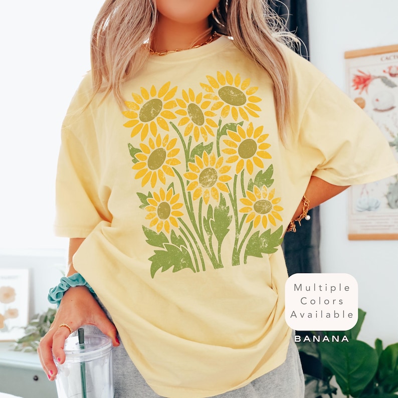 May include: A yellow t-shirt with a vintage-style sunflower print. The text "Multiple Colors Available" and "BANANA" are printed on the bottom right corner of the shirt.