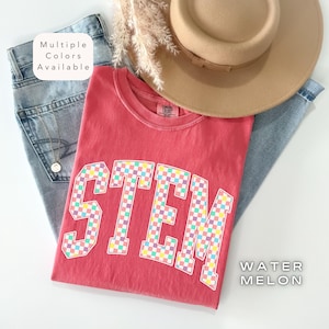 May include: A red t-shirt with the word "STEM" in a checkered pattern, paired with light-wash jeans and a tan hat. The shirt is labeled "Watermelon" and the text "Multiple Colors Available" is also present.