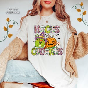 Puede incluir: Camiseta color crema con las palabras "Hocus Crocus" en una fuente colorida y estampada. El diseño presenta una rana de dibujos animados con un sombrero de bruja junto a una calabaza naranja y una seta, con murciélagos. Tema de Halloween.