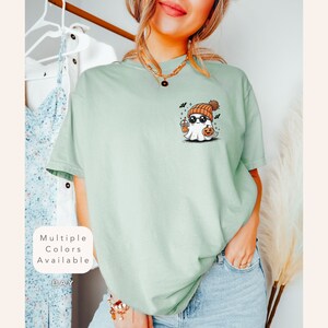 Pode incluir: Uma camiseta verde claro com um fantasma de desenho animado usando um gorro, &oacute;culos de sol e segurando uma ab&oacute;bora e uma bebida. O texto "Multiple Colors Available" est&aacute; no canto inferior esquerdo da imagem.