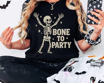 May include: Black t-shirt featuring a skeleton illustration and the phrase "BONE TO PARTY". The skeleton is depicted with raised arms and stars. A Halloween-themed design.