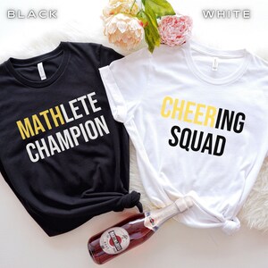Puede incluir: Dos camisetas, una negra y una blanca, con texto impreso en ellas. La camiseta negra dice "MATHLETE CHAMPION" en letras blancas y doradas. La camiseta blanca dice "CHEERING SQUAD" en letras negras y doradas.