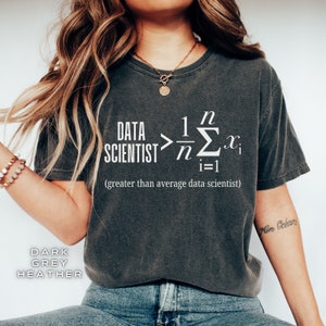 May include: A dark heather grey t-shirt with a white graphic of a data scientist equation. The equation reads "DATA SCIENTIST > 1/n Σ(i=1 to n) x(i)" and the text below the equation reads "(greater than average data scientist)".