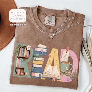 May include: An espresso-coloured t-shirt featuring the word READ in large letters. The R is a bookshelf, the E is a stack of books, the A is an open book, and the D includes a mug with the text Just one more chapter and glasses.