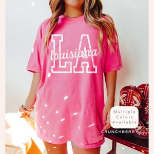 May include: A bright pink t-shirt with the text "LA Louisiana" in white block letters. The shirt is a short-sleeved, crew neck style. The image also shows a pair of red cowboy boots.