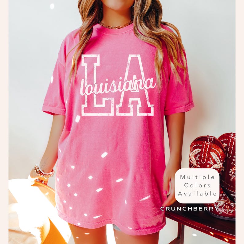 May include: A bright pink t-shirt with the text "LA Louisiana" in white block letters. The shirt is a short-sleeved, crew neck style. The image also shows a pair of red cowboy boots.