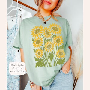 May include: A light blue t-shirt with a vintage-style graphic of sunflowers. The text "Multiple Colors Available" is visible in the lower left corner of the image.