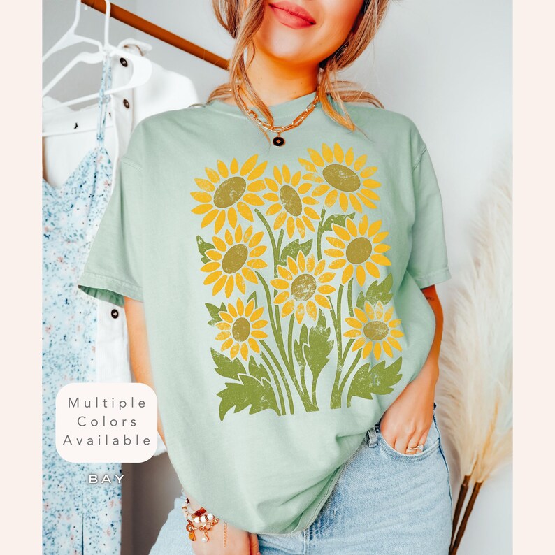 May include: A light blue t-shirt with a vintage-style graphic of sunflowers. The text "Multiple Colors Available" is visible in the lower left corner of the image.