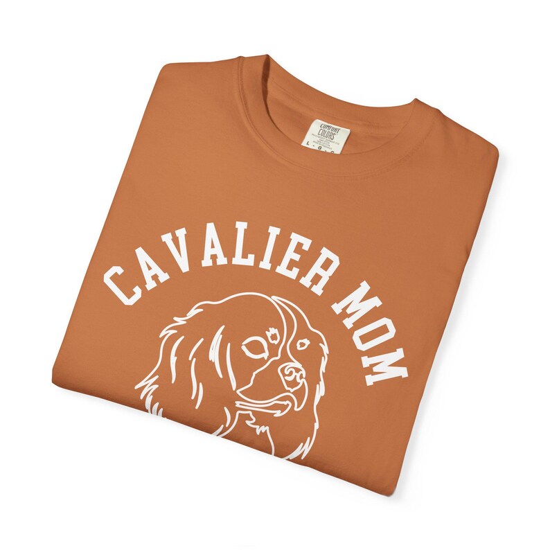 Peut inclure: T-shirt de couleur rouille avec l'inscription blanche "CAVALIER MOM". Un dessin au trait blanc d'un chien Cavalier King Charles Spaniel est centr&eacute; sous le texte. Le t-shirt est pli&eacute;.