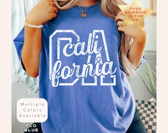 May include: A blue t-shirt with the word "California" in white block letters. The shirt has a distressed, vintage look. The text "Multiple Colors Available" and "FLO BLUE" are also visible.