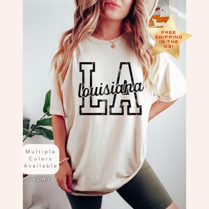 May include: Ivory-colored t-shirt with the text "LA Louisiana LA" in a distressed black font. The shirt has short sleeves and a relaxed fit. The image also includes a gold star with the text "FREE SHIPPING IN THE US!"