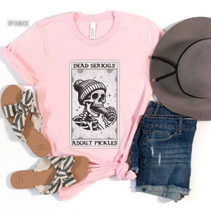 May include: Pink t-shirt with a graphic of a skeleton wearing a beanie and eating a pickle. The graphic is in a card-like frame with the text "Dead Serious About Pickles".