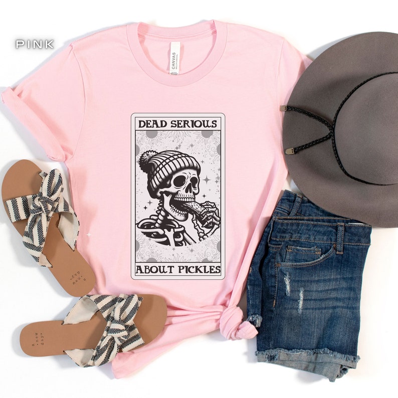 May include: Pink t-shirt with a graphic of a skeleton wearing a beanie and eating a pickle. The graphic is in a card-like frame with the text "Dead Serious About Pickles".